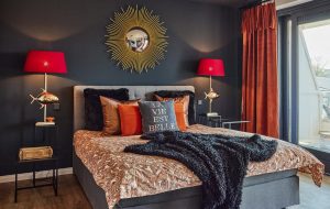 Das Schlafzimmer einer Ferienwohnung mit einer dunklen Wand, goldenem Spiegel, zwei roten Lampenschirmen im Redesign gestaltet