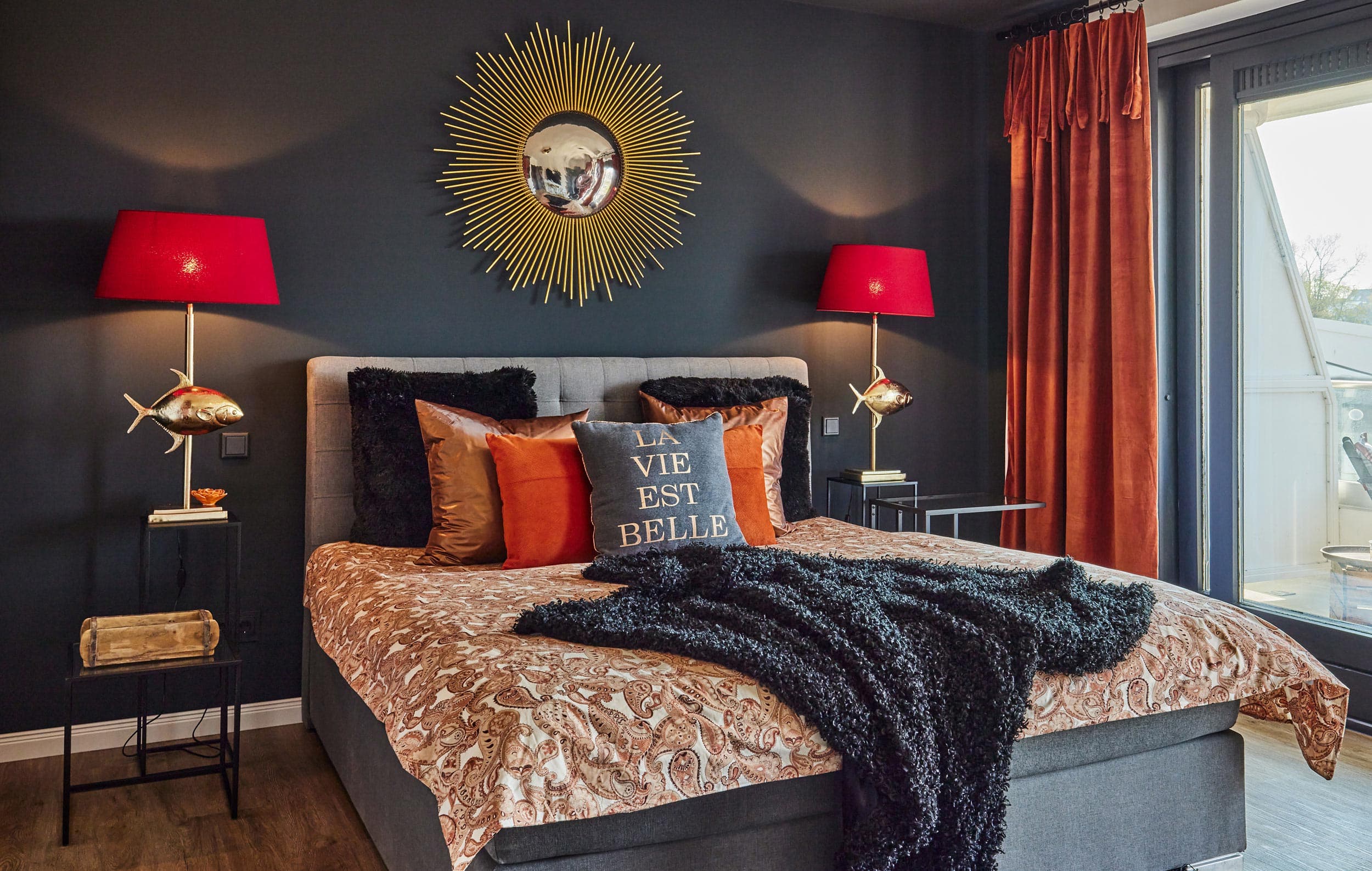 I-LOVE-HOME-Ferienwohnung-Scharbeutz-Schlafzimmerbett_RET Das Schlafzimmer einer Ferienwohnung mit einer dunklen Wand, goldenem Spiegel, zwei roten Lampenschirmen im Redesign gestaltet